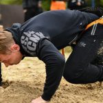 A determined man crawls through sand pulling a rope outdoors, showcasing strength and endurance.